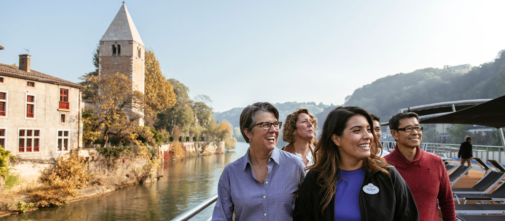 An Adventure Guide and 3 Adventurers pass a stone building and tower as they sail down the Rhône River