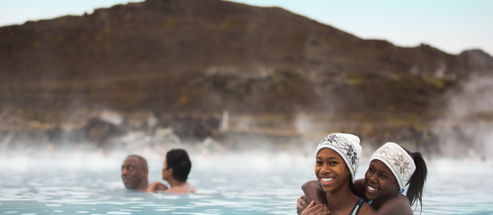 Two girls in matching headbands wade in an outdoor bath of steaming water near a man and a woman with a mountain in the background Two girls in matching headbands wade in an outdoor bath of steaming water near a man and a woman with a mountain in the background
