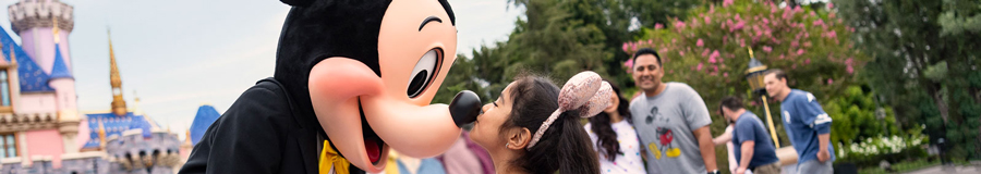 A young girl greets Mickey in front of Sleeping Beauty Castle by kissing him on the nose as he shakes her hands